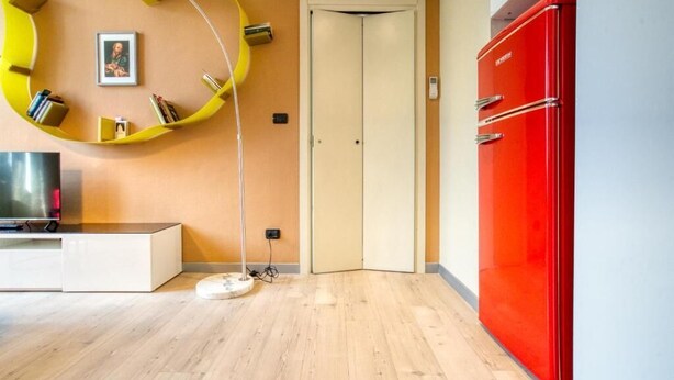 Una habitación con piso de madera, un armario rojo, una estantería circular amarilla y una puerta cerrada, además de un televisor sobre un mueble blanco.