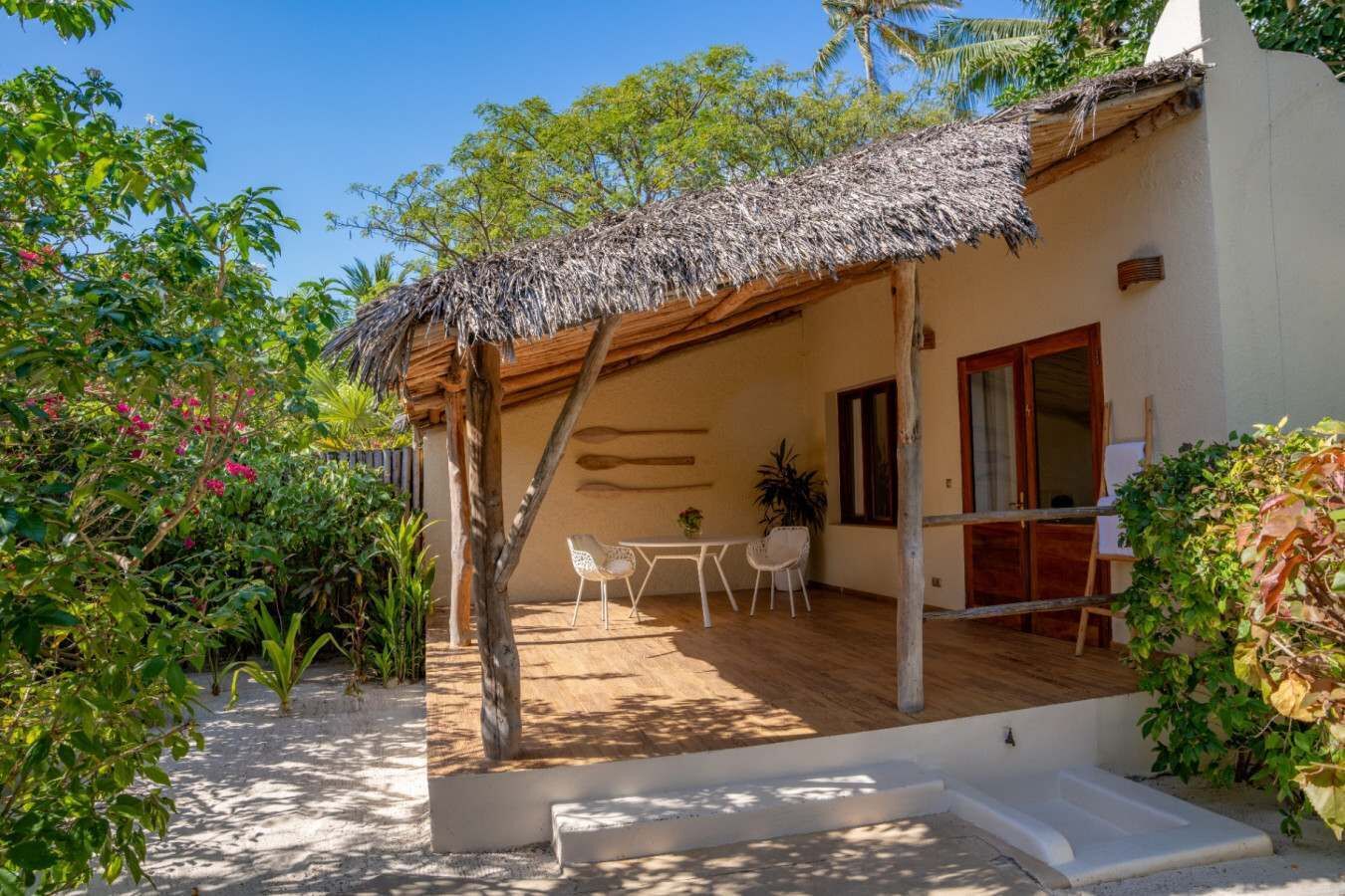 A covered outdoor area with a table and chairs, a thatched roof, and a white building with a door and windows.