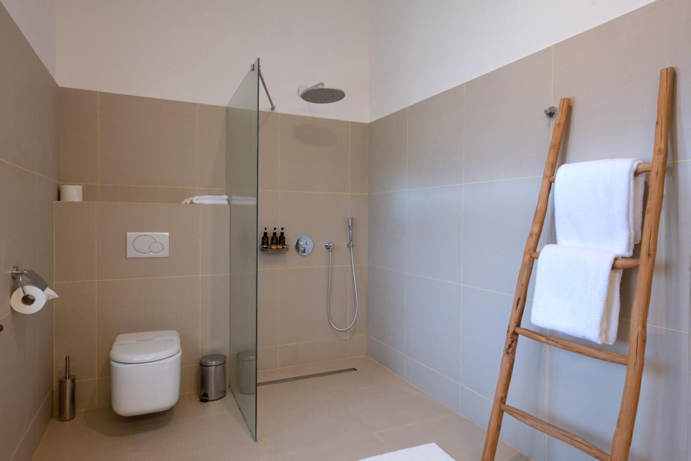 A modern bathroom with a glass shower enclosure, a toilet, a bidet, and a wooden ladder with towels.