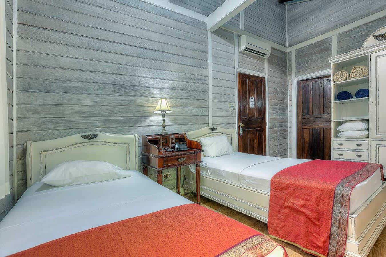 A room with two beds, a nightstand, a wooden wardrobe, and a lamp.