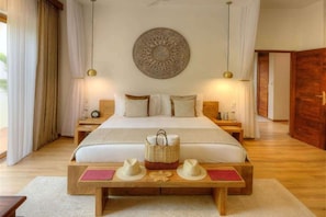A hotel room with a large bed, two bedside tables, a wall-mounted decorative plate, and a wooden bench with hats. - Eco-Luxury Pool Villa (Paje)