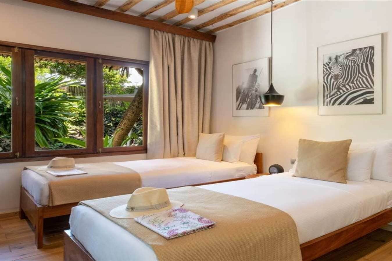 A hotel room with two beds, a window with curtains, a hanging light fixture, and a framed picture on the wall.