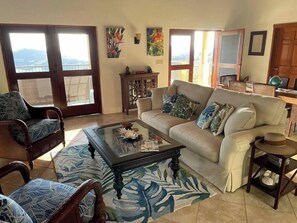 A living room with a sofa, chairs, a coffee table, and a view through a glass door. - Beneath the Mango Tree: Private Pool, Prime Location, Stunning Sunsets! (Cruz Bay)