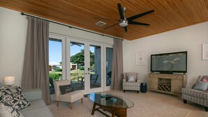 Un salón espacioso con ventilador de techo, una mesa de centro de vidrio, un televisor sobre un soporte de madera y vistas al exterior a través de amplias puertas correderas de cristal.