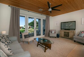 Ein geräumiges Wohnzimmer mit Deckenventilator, einem Glastisch, einem Fernseher auf einem Holzständer und Blick ins Freie durch große Schiebeglastüren.
