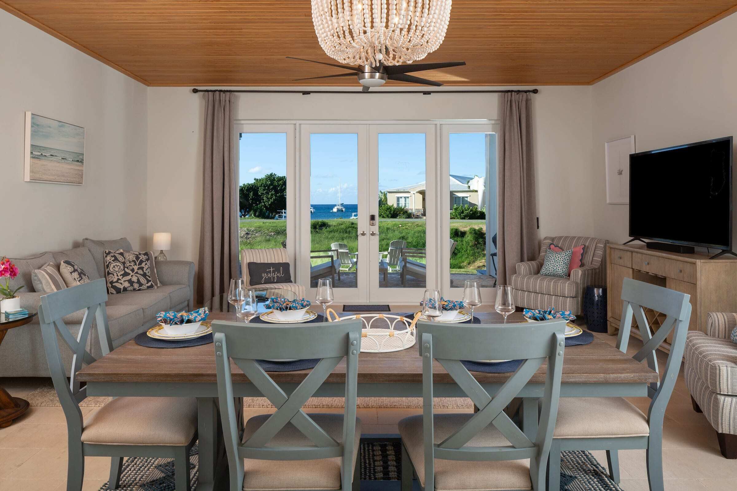 A dining area with a view of the sea through large windows, a chandelier, and a television on the right.