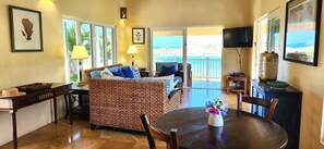 A room with a wicker sofa, a dining table, and a TV. There are windows and a view of the outdoors. - Elysium Villa-Natural Surroundings, Ocean Views, Private Pool! (Cruz Bay)