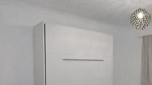 A room with a large white wardrobe, a small round ottoman, a chair, and a hanging light fixture. - Studio Sleeps 2 Near Station & Airport (Bishop's Stortford)