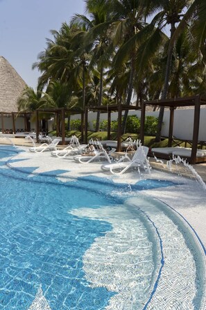 Une piscine d’un bleu éclatant, avec des chaises longues blanches et, au fond, une structure surmontée d’un toit de chaume.