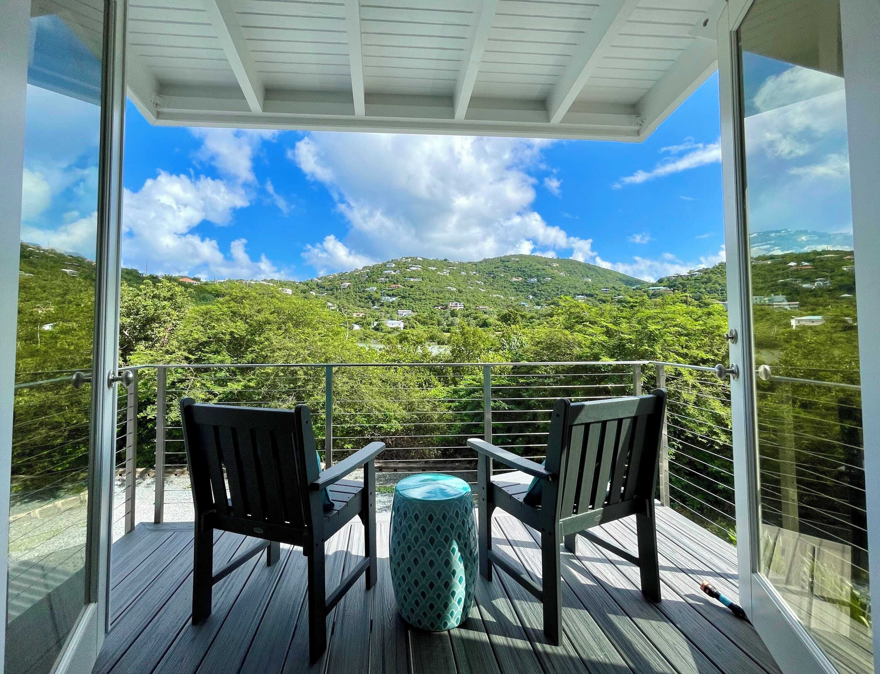 Ein Balkon mit zwei Stühlen und einem kleinen Tisch, der auf eine gebirgige Landschaft mit verstreuten Häusern blickt.