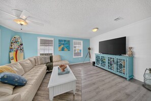 A living room with a beige sofa, a white coffee table, a teal cabinet, a ceiling fan, and a mounted TV. - Beach Breeze Bungalow (Fort Myers)