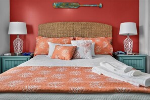 Una cama con cabecera trenzada, varios almohadones con diseño de hojas y una manta con un motivo parecido. Hay dos mesitas de noche con lámparas y objetos decorativos.