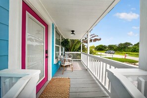 A porch with a white railing, a table with two chairs, a red door, and a view of a road and greenery. - Beach Breeze Bungalow (Fort Myers)
