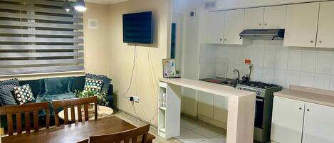 A compact kitchen with a dining area, a TV mounted on the wall, and a window with blinds.