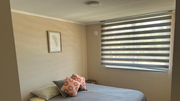 Un dormitorio con cama, ventana con persianas, un cuadro en la pared y una mesita de noche.
