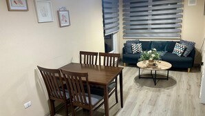 Un comedor con mesa y sillas de madera, una mesita de centro redonda con un florero, un sofá azul oscuro con cojines estampados, y una ventana con persianas.