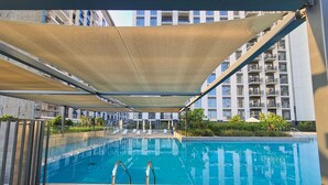 A swimming pool with a shaded area, surrounded by modern buildings. - Free Housekeeping Visits During Your Stay - Stayshort - Chic 1-bed for 4 in Prime Dubai Hills Location (Dubai)