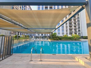 A swimming pool with a shaded area, surrounded by modern buildings. - Free Housekeeping Visits During Your Stay - Stayshort - Chic 1-bed for 4 in Prime Dubai Hills Location (Dubai)