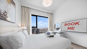 A hotel room with a large bed, a view of the city, a 'BOOK ME' sign, and a desk with a lamp. - Free Housekeeping Visits During Your Stay - Stayshort - Chic 1-bed for 4 in Prime Dubai Hills Location (Dubai)