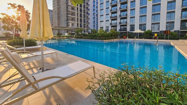 A swimming pool with lounge chairs and umbrellas, surrounded by greenery and modern buildings. - Free Housekeeping Visits During Your Stay - Stayshort - Chic 1-bed for 4 in Prime Dubai Hills Location (Dubai)