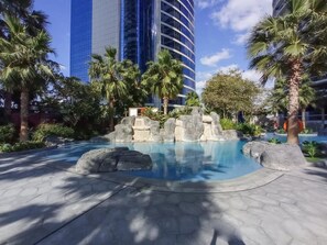 A swimming pool with rock formations, surrounded by palm trees and a modern building in the background. - Free Housekeeping Visits During Your Stay - Stayshort - Modern 2BR in Prime Business Bay 4 Guests (Dubai)