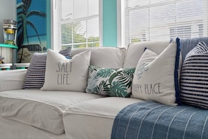 A white sofa with multiple cushions, some with text, in a room with a window and a painting. - 3BR/2BA Beach House-Tiki Bar! King ensuite w/ deck (Fort Myers)
