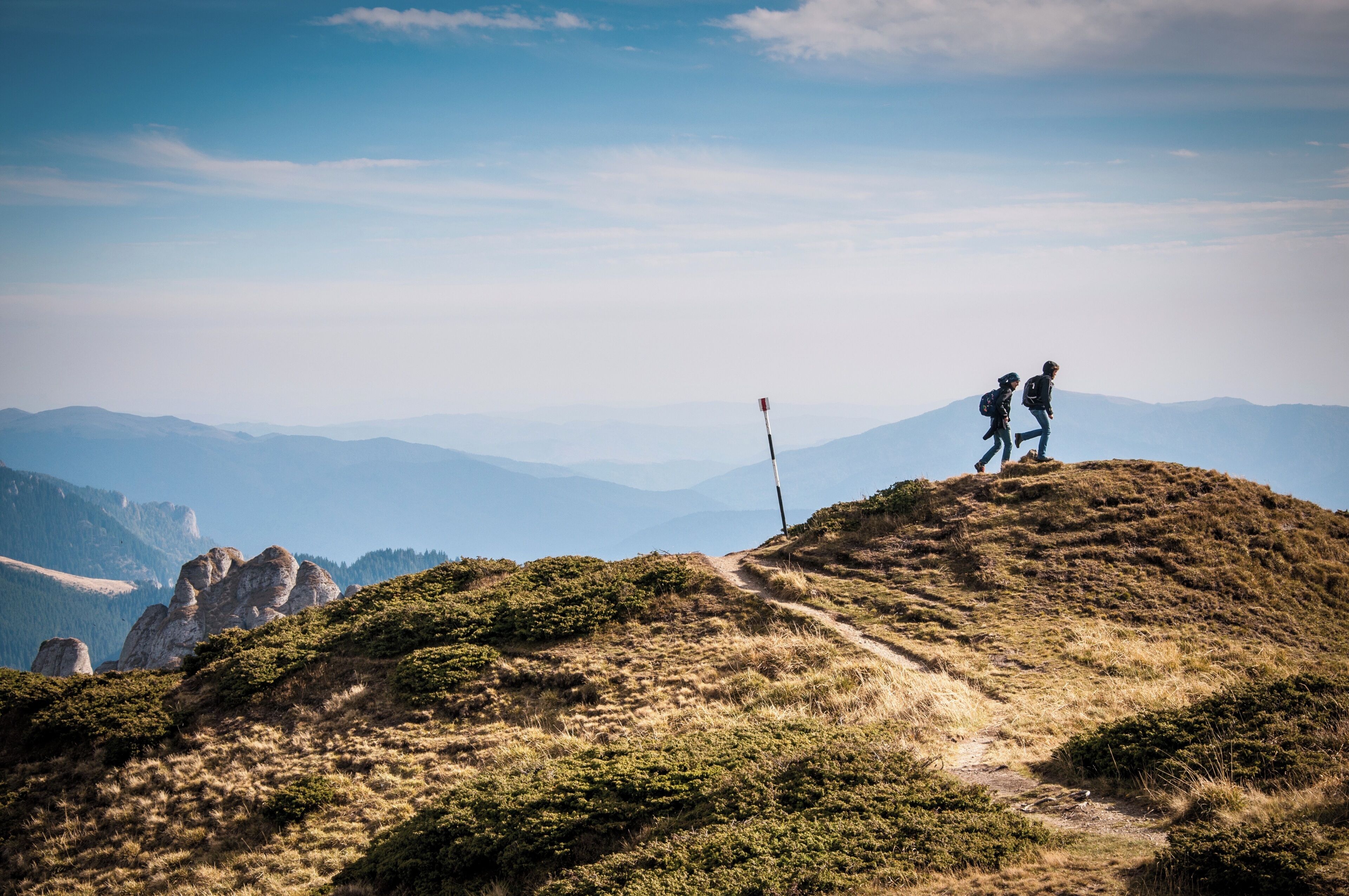 A hiker's path on a grassy hill with a flag marker, surrounded by mountains.