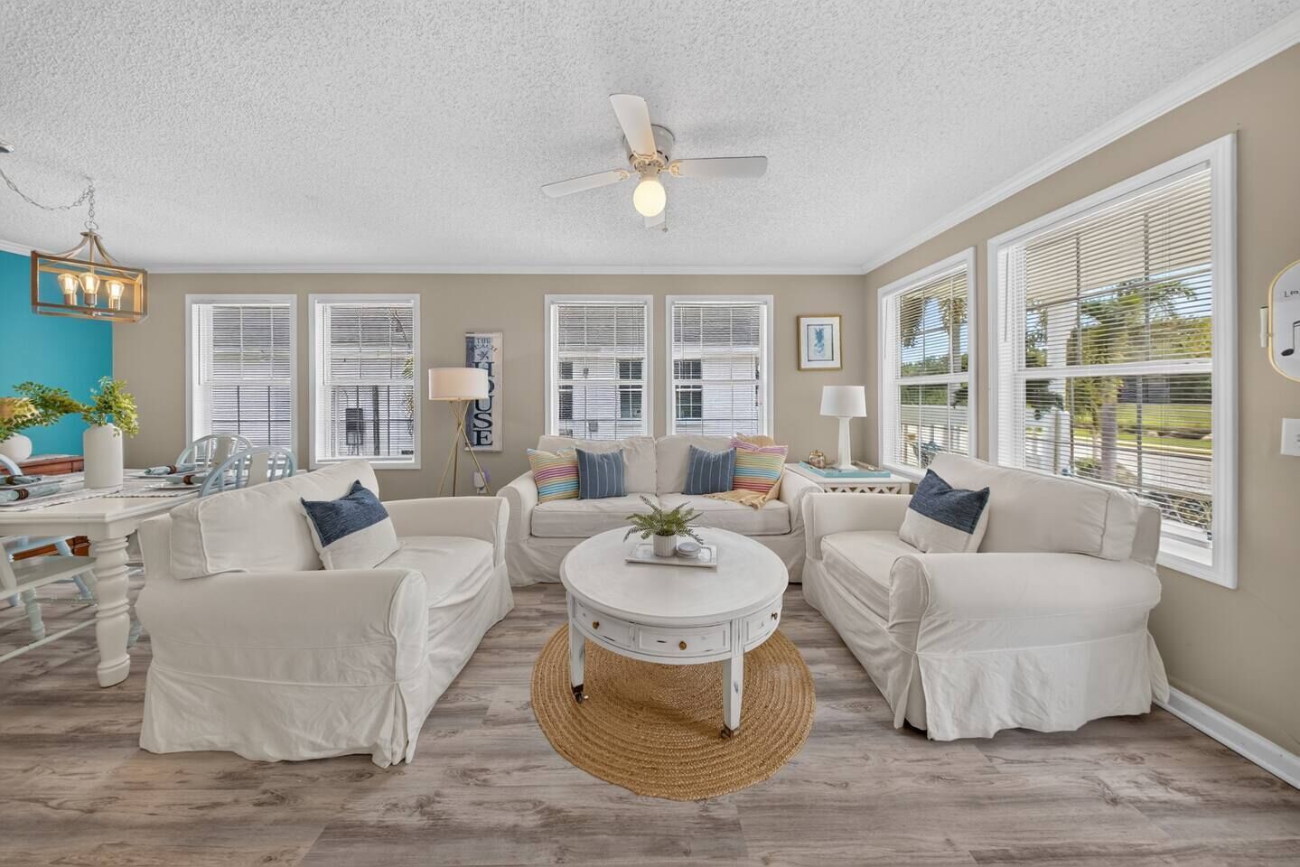 A spacious living room with a ceiling fan, large windows with blinds, and a central coffee table.