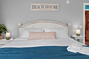 Ein ordentlich bezogenes Bett mit einem karierten Kissen, eine Nachttischlampe und ein Wandschild mit der Aufschrift 'BEACH HOUSE FORT MYERS BEACH, FL'.