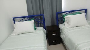Een kamer met twee eenpersoonsbedden, elk met een blauw metalen frame en witte beddengoed. Tussen de bedden staat een klein zwart nachtkastje met lades.