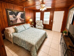 Ein Schlafzimmer mit einem Bett, einem Sessel, einem Deckenventilator, einem Gemälde und einer Tür.