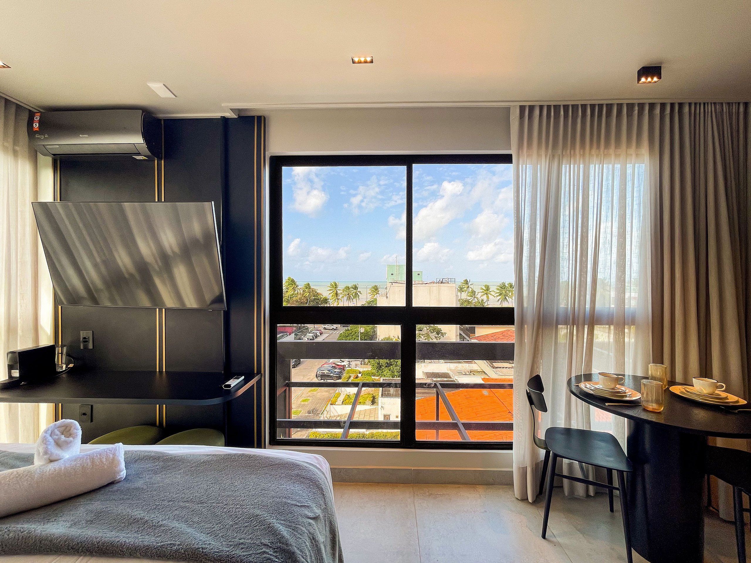 A hotel room with a bed, a desk, a chair, a table with a cup and saucer, a window with a view of the city, and an air conditioning unit.