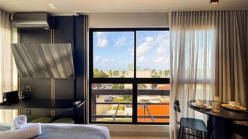 A hotel room with a bed, a desk, a chair, a table with a cup and saucer, a window with a view of the city, and an air conditioning unit.