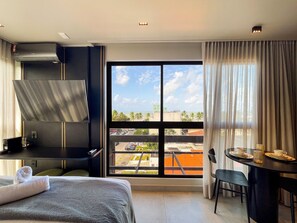 A hotel room with a bed, a desk, a chair, a table with a cup and saucer, a window with a view of the city, and an air conditioning unit.