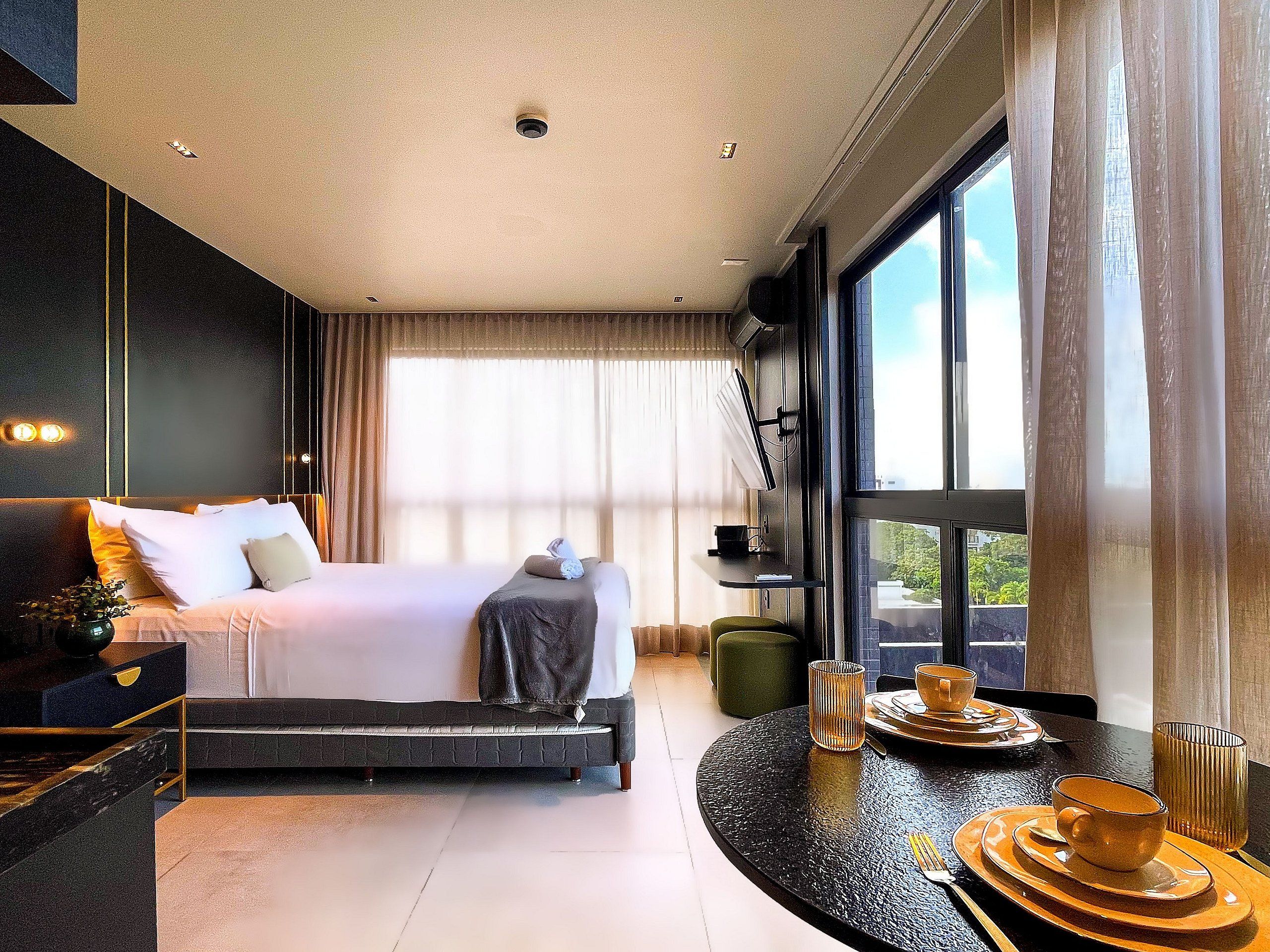 A modern hotel room with a large bed, a seating area with a table, and a view of the outdoors through large windows.