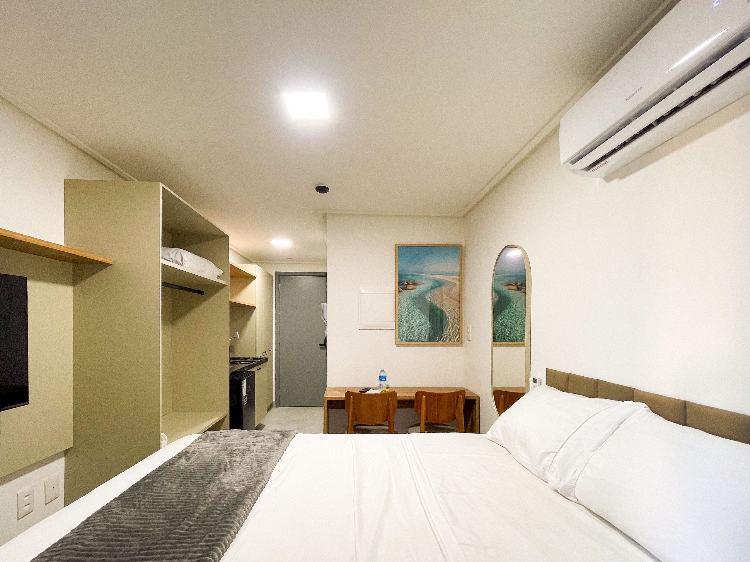 Quarto de hotel com uma cama, uma mesa, uma cadeira, um espelho e um quadro de uma praia.