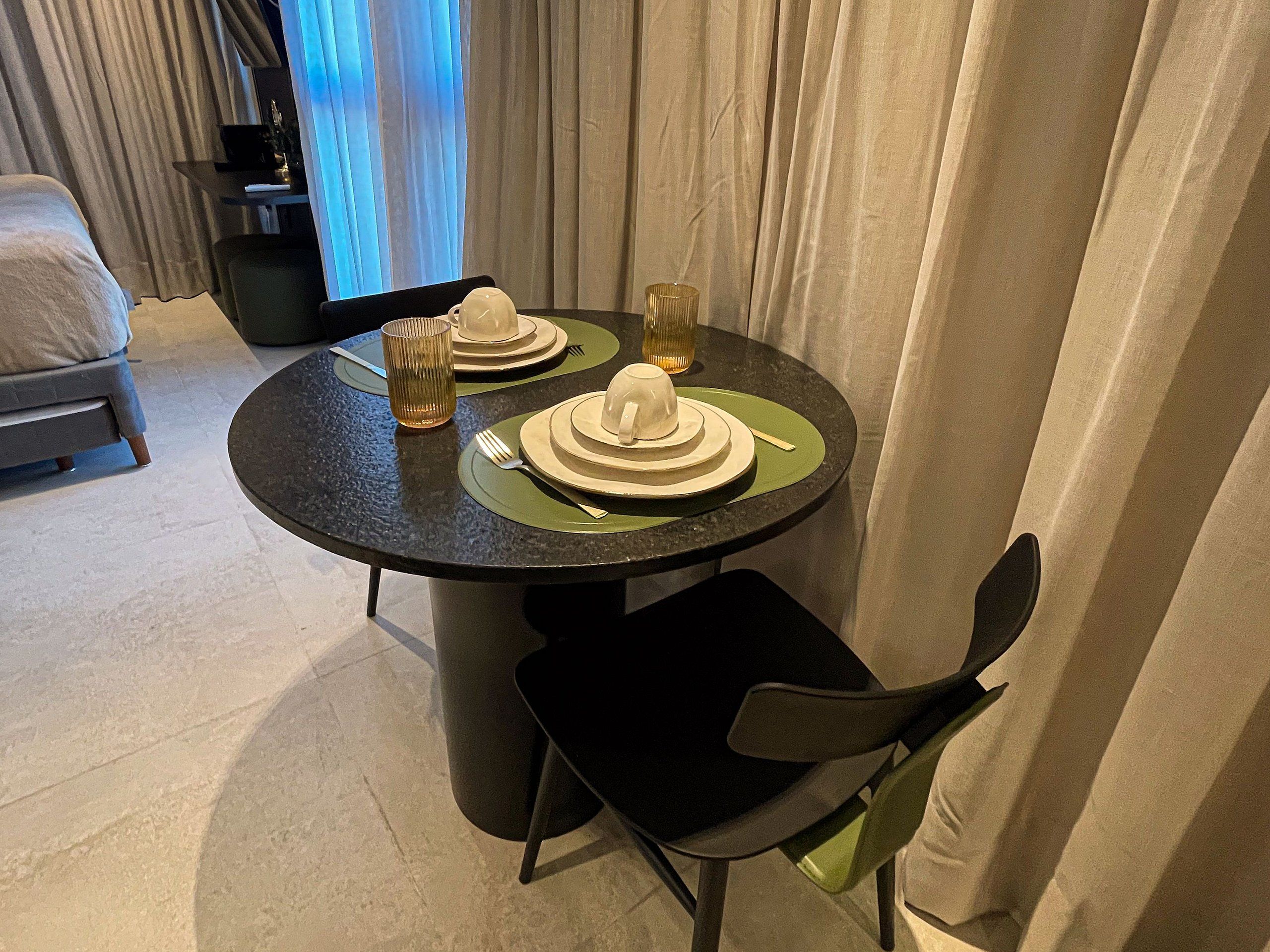 Mesa de jantar posta com pratos, copos e talheres, uma cadeira e uma cama ao fundo.