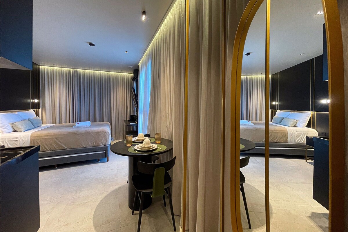 A modern hotel room with a large bed, a small dining table, and a mirror reflecting the room.