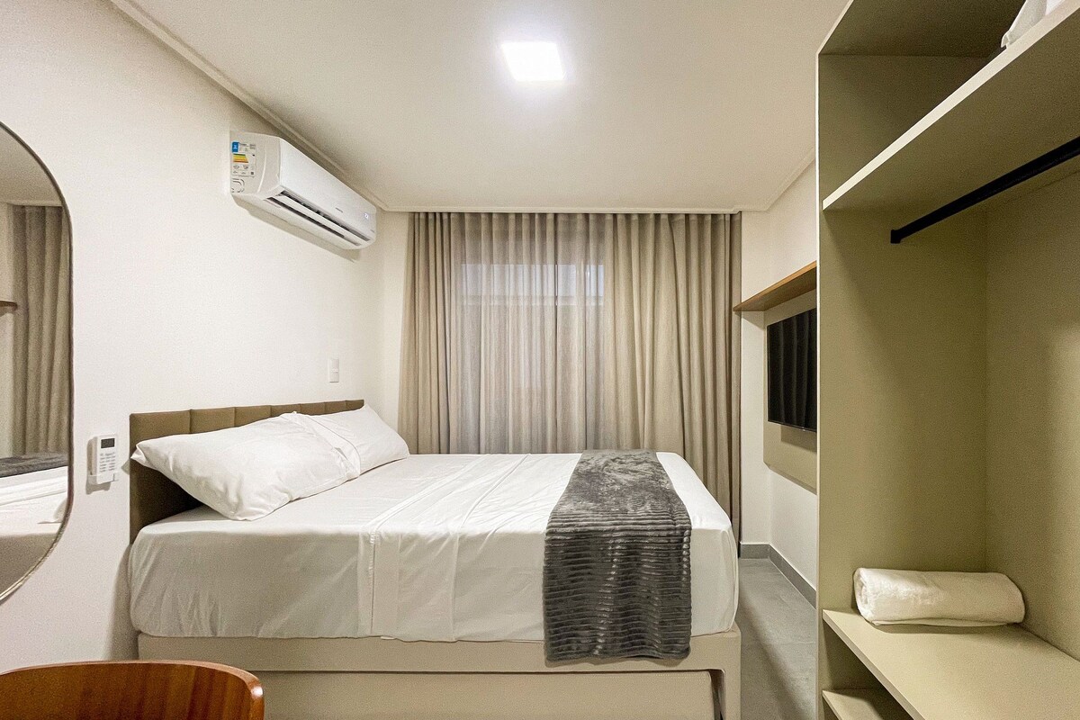 A hotel room with a bed, a bedside table, a chair, a wardrobe, and an air conditioning unit.