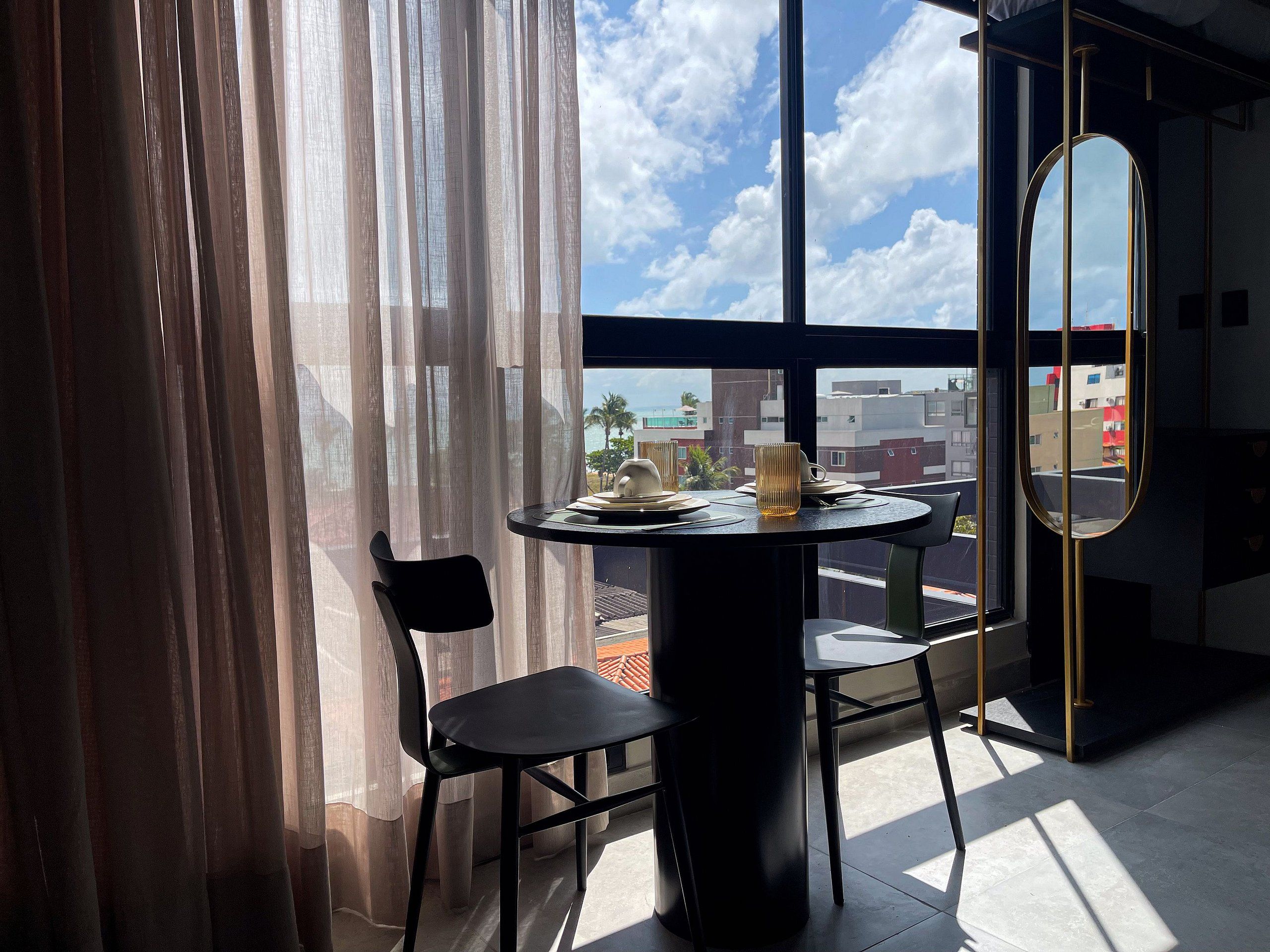 Una habitación con una mesa redonda, una silla, un ventanal con vistas a edificios y palmeras, y un espejo en la pared.