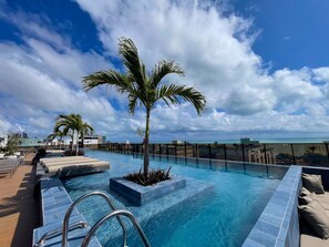 Piscina com palmeira, espreguiçadeiras e vista para o oceano.