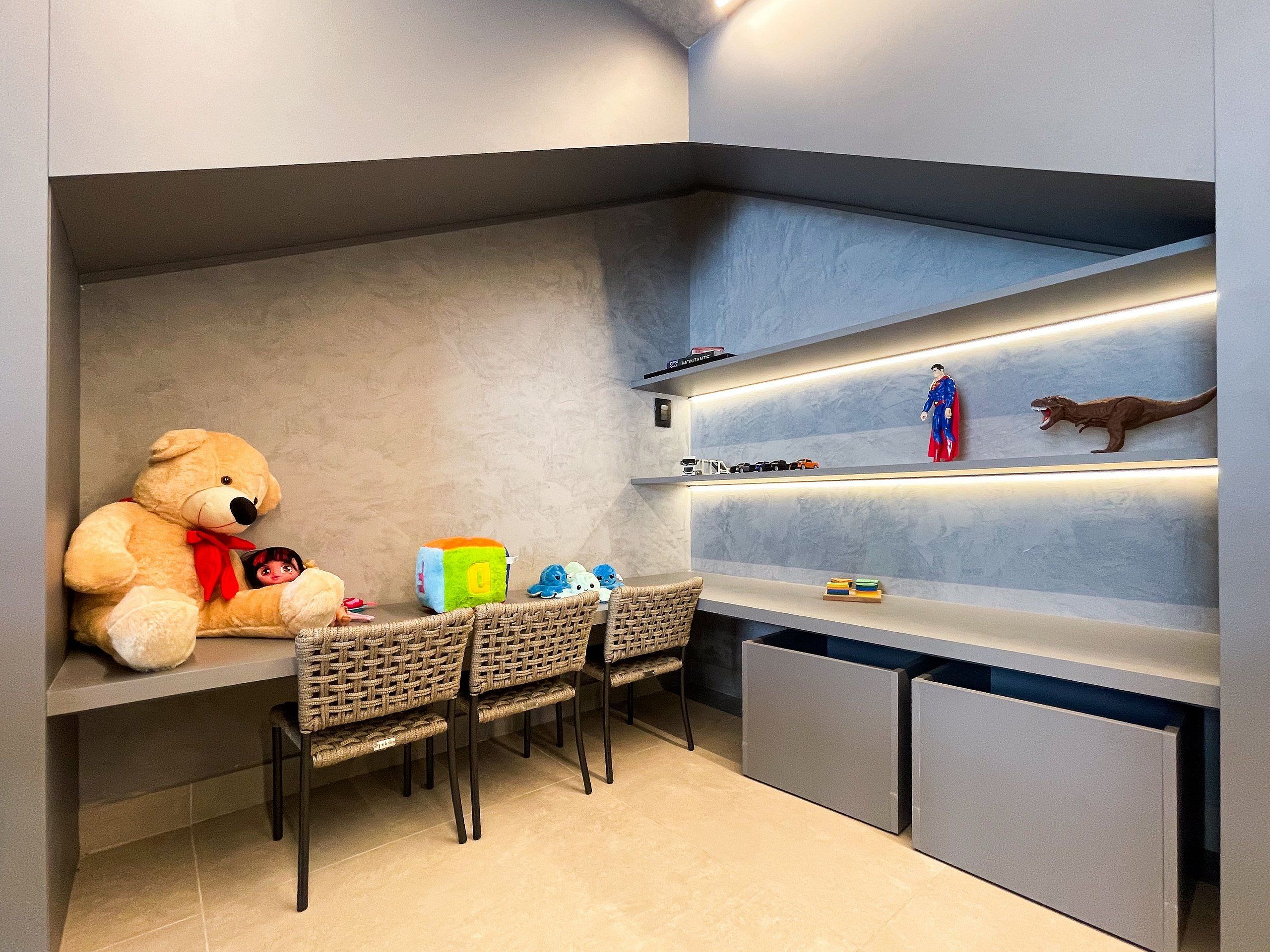 Espaço de recreação infantil com um urso de pelúcia grande, uma mesa pequena, cadeiras e prateleiras com brinquedos e figuras.