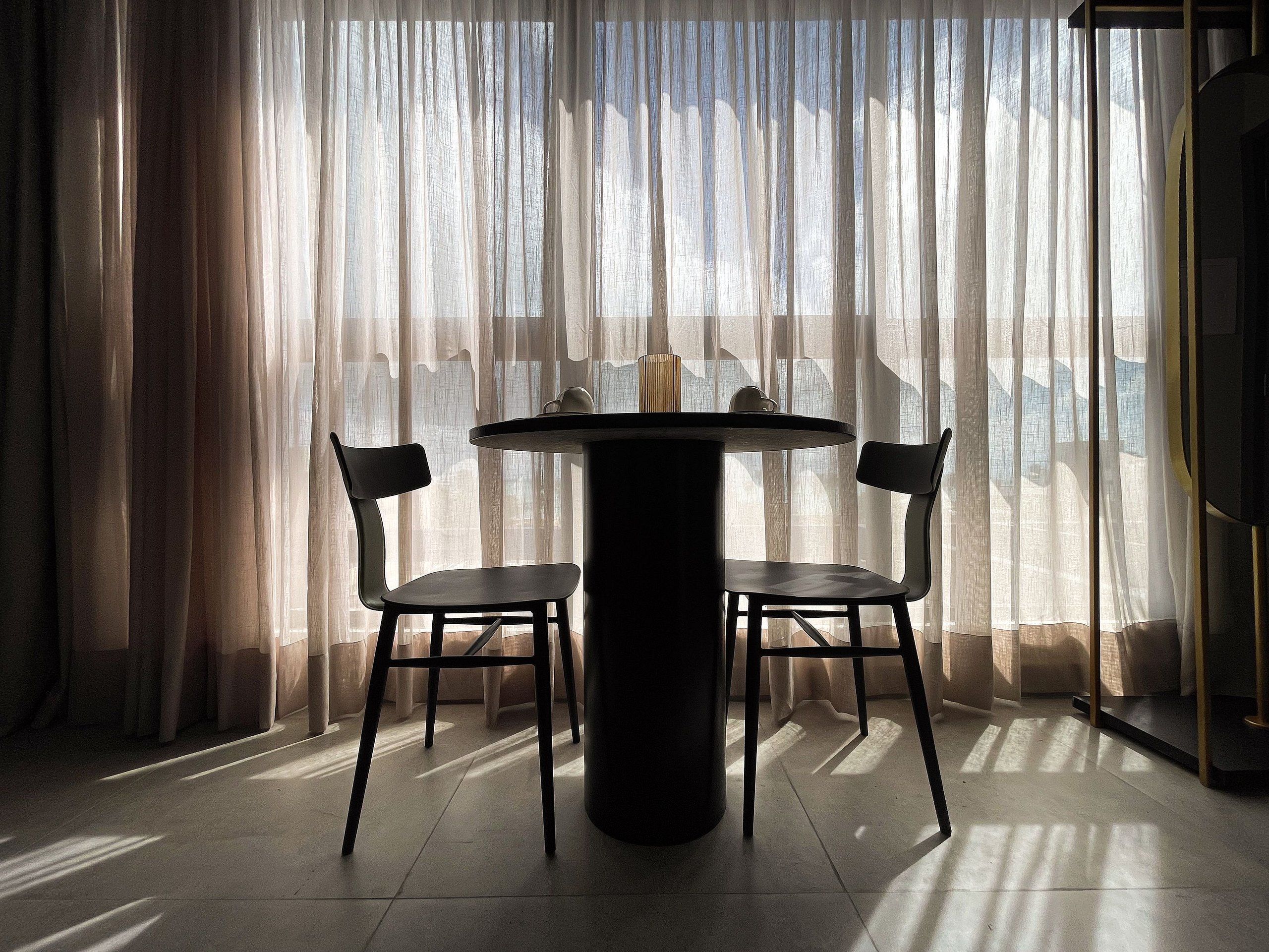 Quarto com mesa redonda e duas cadeiras, janela com cortinas leves e vista para o exterior.
