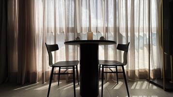 Quarto com mesa redonda e duas cadeiras, janela com cortinas leves e vista para o exterior.