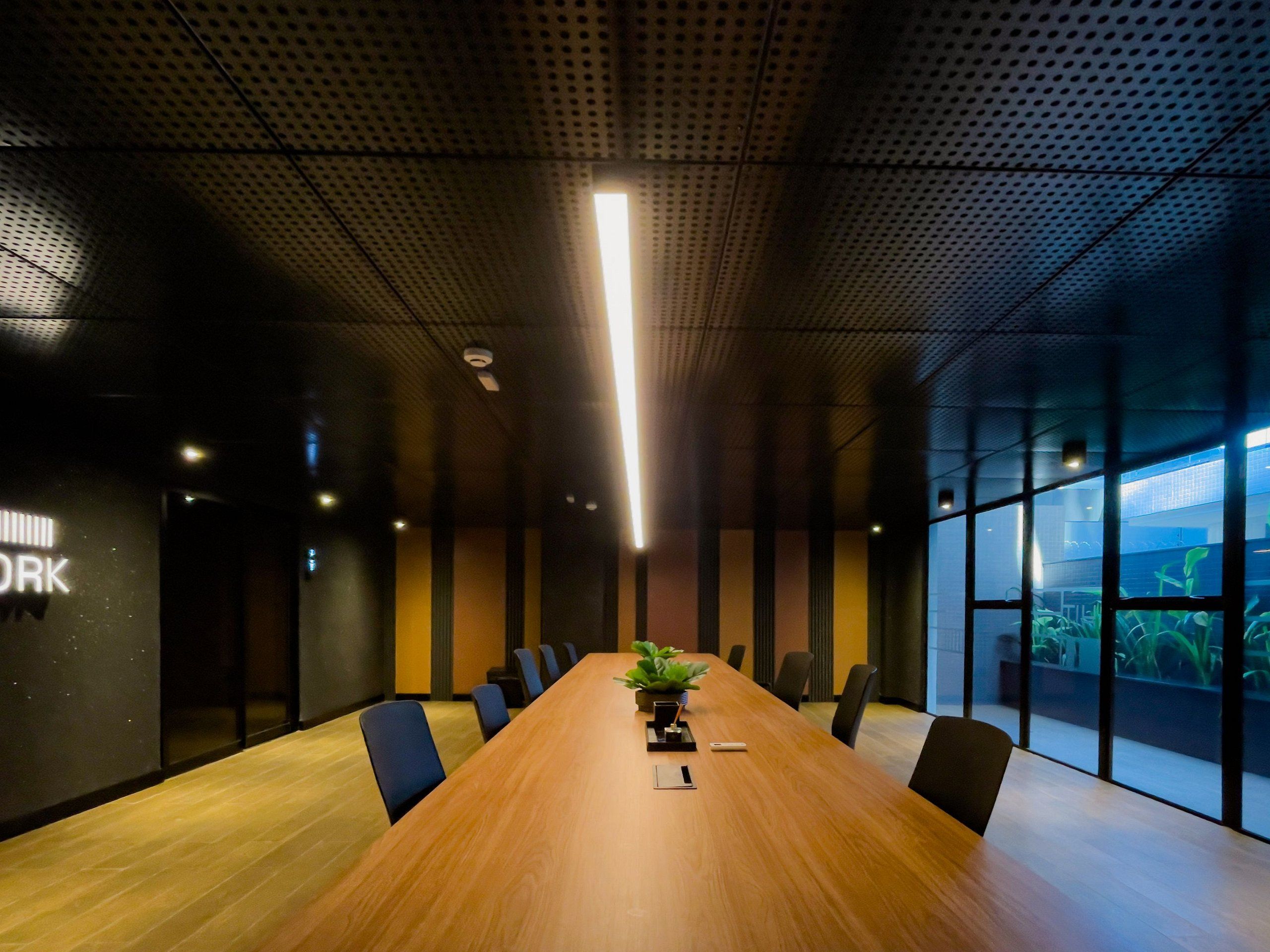 Un salón de conferencias con una larga mesa de madera, sillas negras, una planta en maceta y un amplio ventanal con vistas a zonas verdes.