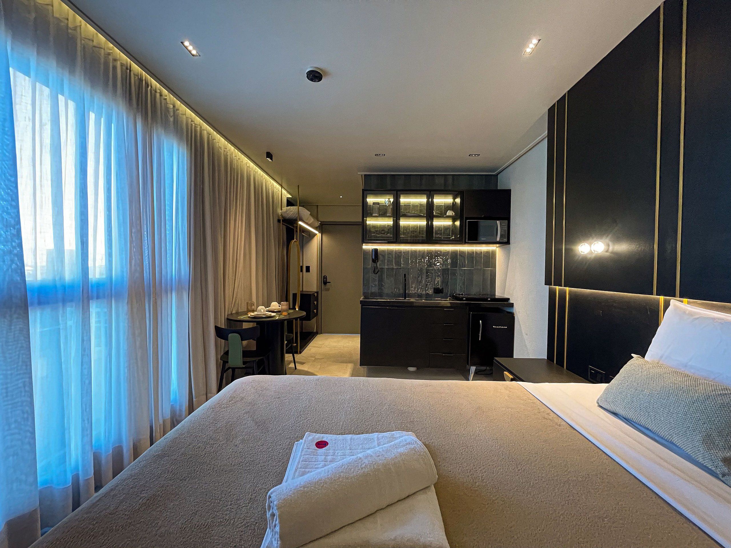 A modern hotel room with a large bed, a desk, and a kitchenette.