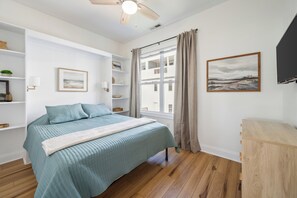 A bedroom with a bed, a window with curtains, a ceiling fan, and a painting on the wall. - Salt & Sand on Bowfin (Carolina Beach)