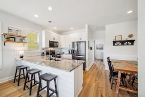A modern kitchen with white cabinetry, a granite countertop, and stainless steel appliances. There are bar stools and a dining table with chairs. - Salt & Sand on Bowfin (Carolina Beach)