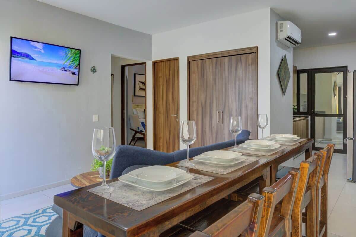 A dining area with a long wooden table set for four, flanked by two wooden doors, a television mounted on the wall, and an air conditioning unit.