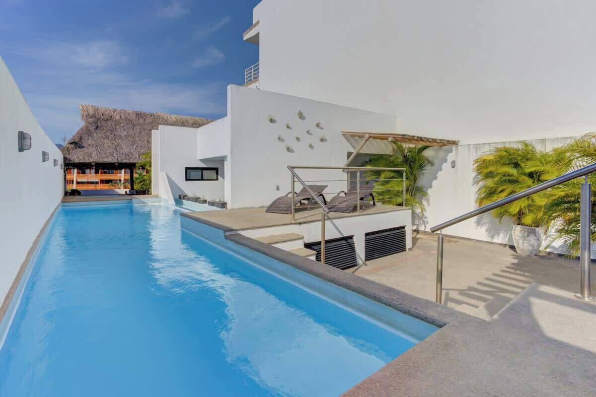 A swimming pool with a sun terrace, a thatched-roof structure, and palm trees.